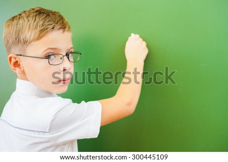 First grade schoolboy wrote on blackboard with chalk at classroom and looking at the camera. Little boy dressed in a white shirt and wearing glasses. Concept of knowledge day and first of september