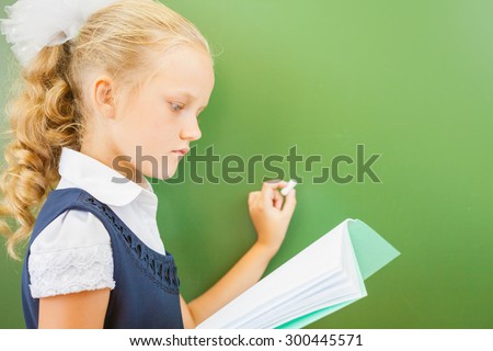 First grade schoolgirl holding a notebook and wrote on blackboard with chalk at classroom. Little girl dressed in a school uniform. Concept of knowledge day and first of september