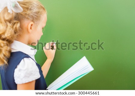 First grade schoolgirl holding a notebook and wrote on blackboard with chalk at classroom. Little girl dressed in a school uniform. Concept of knowledge day and first of september
