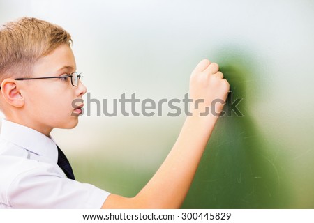 First grade schoolboy wrote on blackboard with chalk at classroom. Little boy dressed in a white shirt and wearing glasses. Concept of knowledge day and first of september.