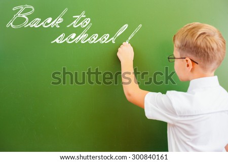 First grade schoolboy wrote on blackboard with chalk - Back to school, at classroom. Left-handed little boy dressed in white shirt and wearing glasses. Concept of knowledge day and first of september