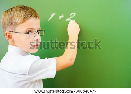 First grade schoolboy wrote on blackboard with chalk - arithmetic digits One Plus Two, and looking at the camera. Boy dressed in a white shirt and wearing glasses. Concept of knowledge day