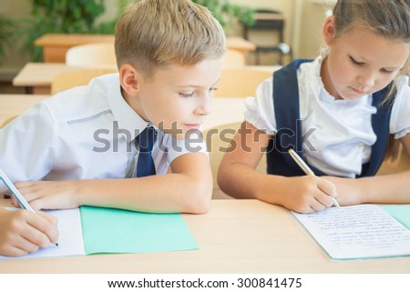 Students or classmates in school classroom sitting together at desk. Boy writes off test at girl, he looking at her notebook. They are dressed in school uniforms. On table there is notebook and pen.