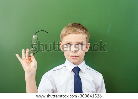 Happy schoolboy standing near the blackboard in a school classroom, dressed in a school uniform, and holding medical glasses. He looking up. There is concept or idea copy space