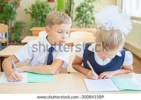 Students or classmates in school classroom sitting together at desk. Boy writes off test at girl, he looking at her notebook. They are dressed in school uniforms. On table there is notebook and pen.