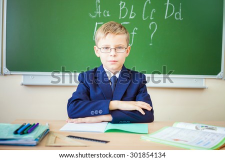 Boy thinking and sits at a desk at school with glasses. Against the background of the school board english alphabet. Schoolboy dressed in a school uniform.