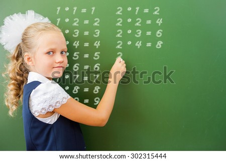 First grade happy schoolgirl wrote on blackboard multiplication table with chalk at classroom. Little girl dressed in a school uniform. Concept of knowledge day and first of september