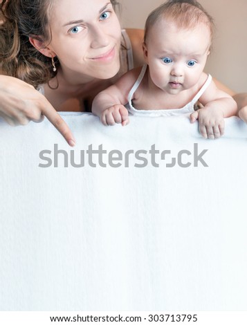 Happy mother and baby lying on a white blanket. Advertising banner sign - Mom is pointing down and surprised baby looking down on empty blank billboard or sign board. Copy space for text and design