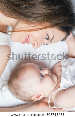 Happy baby girl lying near her mother on a white bed. Girls looking at each other. Mother care is most important in baby life