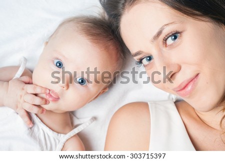 Happy baby girl lying near her mother on a white bed. Newborn looking at the camera and smiling. Child biting her finger, because teeth grow and itching gums.