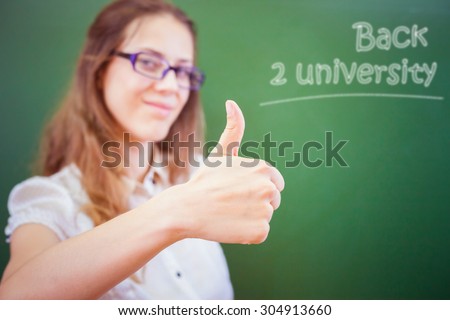 Successful happy young teacher or student at university or school classroom near a blackboard and show a thumbs up gesture. Concept of back to university. Selective focus on the hand