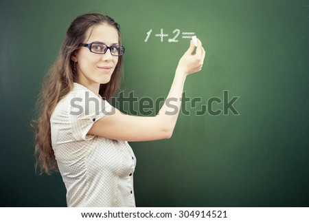 Teacher or student wrote on blackboard with chalk - arithmetic digits one plus two, and looking at the camera. Girl dressed in a white shirt and wearing glasses. Concept of school education