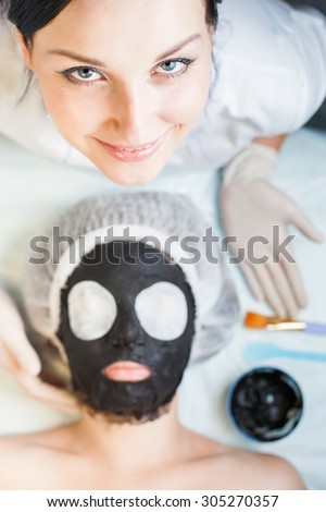Professional woman, cosmetologist in spa salon applying mud face mask. She is happy and looking up at camera. Concept of beauty, healthy therapy, rejuvenation, skincare and relaxing at luxury resort
