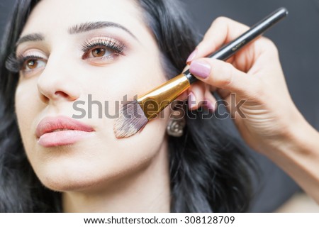 Beautiful arabian woman at beauty salon with a nice makeup. Make up artist apply foundation cream on face, holding in hands a makeup brushe on a dark or black background.