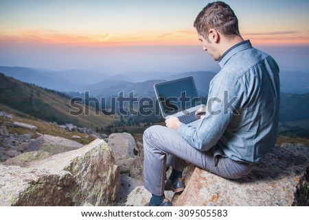 Image of a businessman on the top of the mountain, using a laptop. At the background there are beautiful panorama of valley with high peaks and beautiful sunset