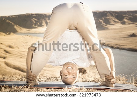 Retro revival image of funny man doing exercise of yoga outdoor above river valley, dressed in casual wear. He stretching and standing like inverted pose.