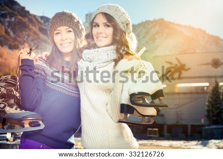 Happy multiracial women going to ice skating outdoor. Dressed in pullover and warm hat. Holding skates shoes. Healthy lifestyle and winter sport concept at sports stadium, mountain landscape