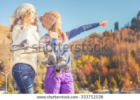 Happy multiracial women pointing on skate rink. Going to ice skating outdoor. Holding skates shoes. Healthy lifestyle and winter sport concept at sports stadium, mountain landscape