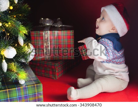 Happy baby surprised near the decorated Christmas tree with many gift and present box! Kid dressed in red Santa hat. Xmas and New Year holiday!