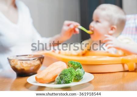 Mom feeds the baby soup. Healthy and natural baby food. Vegetables, carrots, cabbage, broccoli. Child sitting on the highchair at the table.