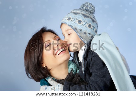Happy family in winter clothing. Little son kissing his happy smiling mother on a cheek over blue