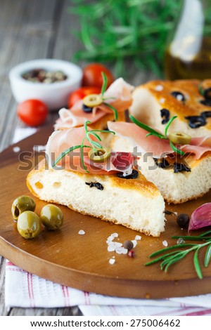 Freshly baked traditional Italian focaccia bread with rosemary, prosciutto ham and black olives on old wooden background, selective focus