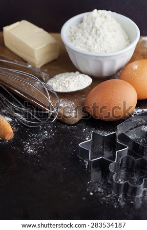 Baking cake ingredients with raw eggs, rolling pin, flour and cookie cutters on black background, selective focus