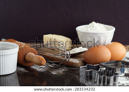 Baking cake ingredients with raw eggs, rolling pin, flour and cookie cutters on black background, selective focus