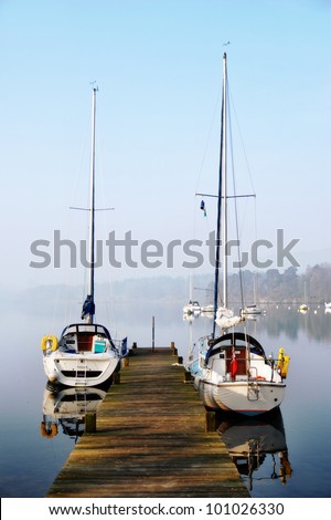 Two small pleasure yachts moored alongside a rustic wooden jetty with mist hanging over the water in Windermere, English Lake District