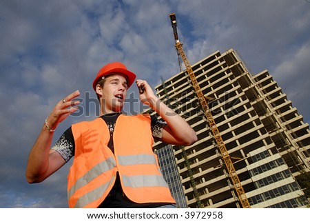 Construction supervisor in safety helmet and reflex vest talking on the phone in front of construction site.