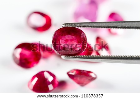 Ruby gemstone held by tweezers. Pile of rubies out of focus in background.