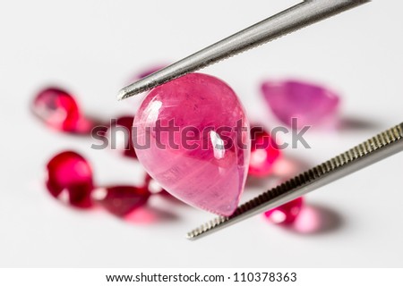Ruby gemstone held by tweezers. Pile of rubies out of focus in background.