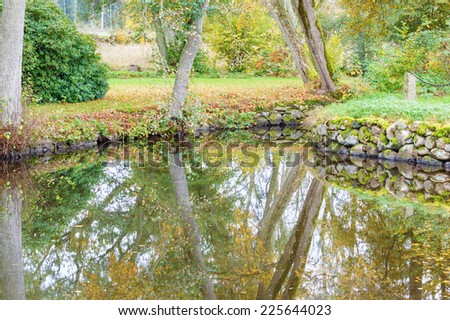 Old pond in garden. Granite boulders used to keep water from running of. Autumn with fallen leaves on ground. Very still water with mirror like reflections.