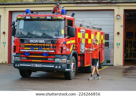 RONNEBY, SWEDEN - OCTOBER 26, 2014: Firefighter in shorts and tshirt walking to fire truck outside station building. Truck is red and yellow Scania 114G.