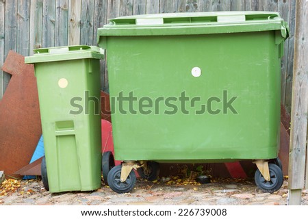 Two green garbage bins with junk behind, Both are closed and have wheels for easier transport. One is big and the other small.