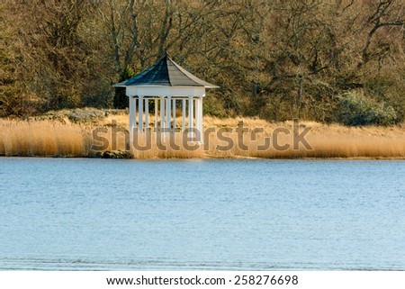White open gazebo by the water in early spring. Reeds surrounding building and trees in background.