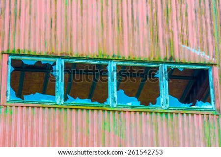 Four broken windows in a row on old industrial building. Wall is metal with red paint and lots of green moss. Blue sky reflected in glass. Inside roof visible in holes.