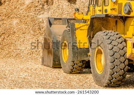 Yellow front loader with bucket down scooping wood chips for biofuel. View from left behind vehicle with pile in front of it.