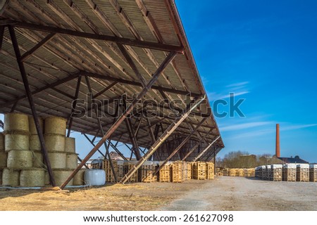 Long roof protecting straw bales and fire wood. To the right is factory smoke stack and more fire wood in pallets. Dirt road follow roof into distance.
