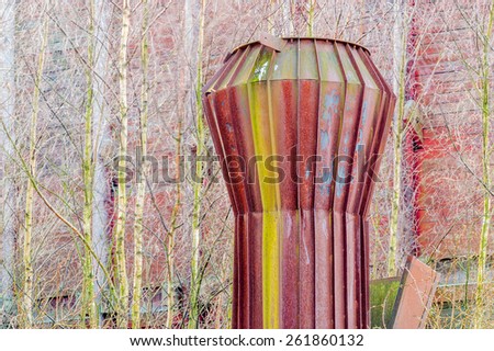 Part of an old boiler chimney or smoke stack that is full of rust and algae. Trees and building in background. Bright red rusty color.