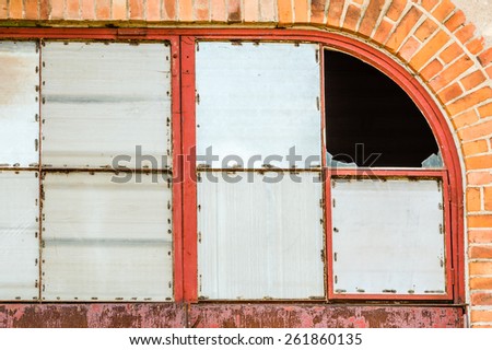 Windows covered with metal on red brick wall. One corner window is broken with glass still present. Darkness inside building.