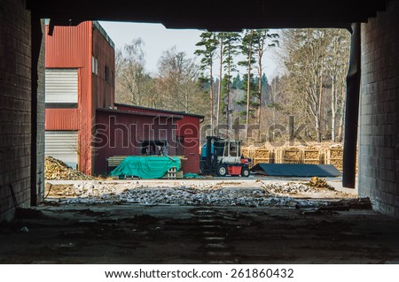 Fire wood industry seen from inside abandoned building. Walls frame the view. Fork lift and fire wood on pallets outside red building. Pile of timber under green tarp in front of building.