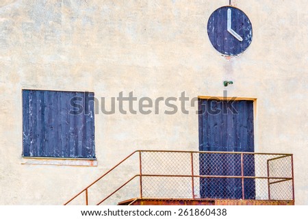 Door and window covered with black painted planks on old beige industrial building. Clock on wall fake made of black painted planks and wooden pointers. Rusty stair with mesh fence leads up to door.