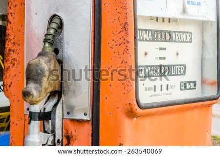 Old vintage gasoline pump seen from side with handle to its side. Orange painted metal with rust and window showing price and volume in white.