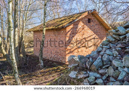 Small red brick building in the forest close to pile of stones. Birch trees grow close to house.
