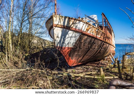The front end of an old passenger ship, now wrecked and pulled on shore. Nature is closing in. Fine blue sky and vegetation all around.