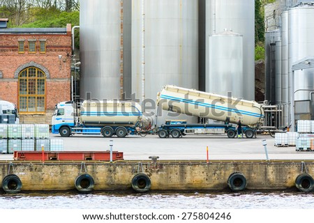 KARLSHAMN, SWEDEN - MAY 06, 2015: Unknown worker standing by trailer and empty his cargo into big silos in background. Seen from across the harbor with water in foreground.