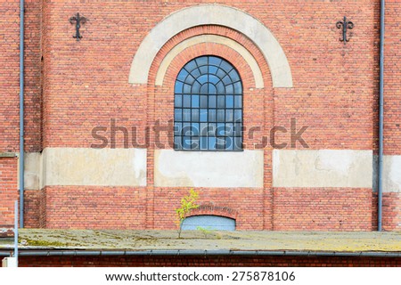 Old brick building in an industrial area. One fine glass window with stone arch over it. Small tree start to grow on roof of building under it.