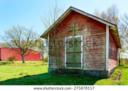 Small red and weathered house without windows. Closed green door and a wooden cross over it. Modern red metal building in background. Used by military, perhaps as church?