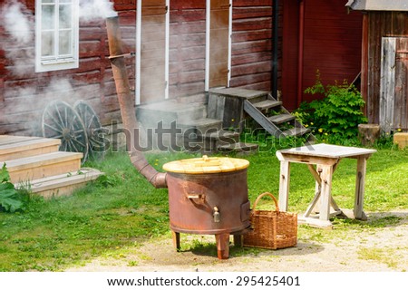 Boiling water in large stove for outdoor laundry. Smoke come out of chimney. Red wooden house in background. This is how it was done in the old times.